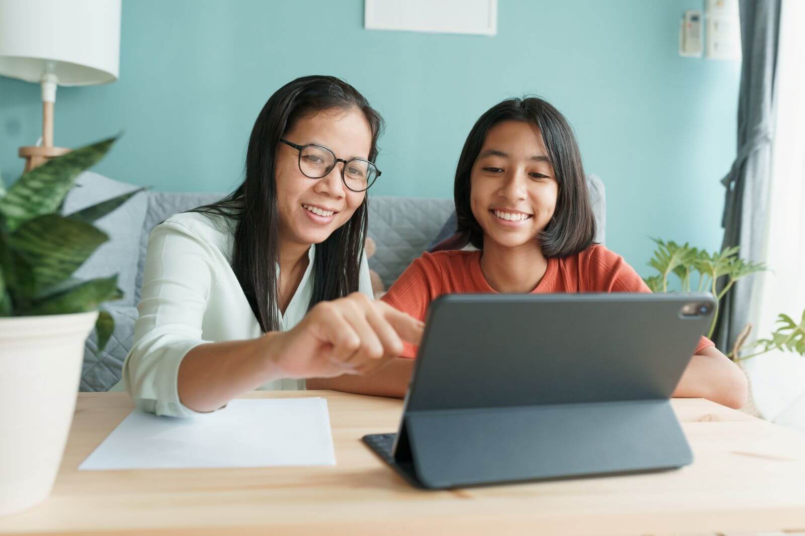 Smiling mother and daughter using a tablet together at a light wooden table with a houseplant nearby.