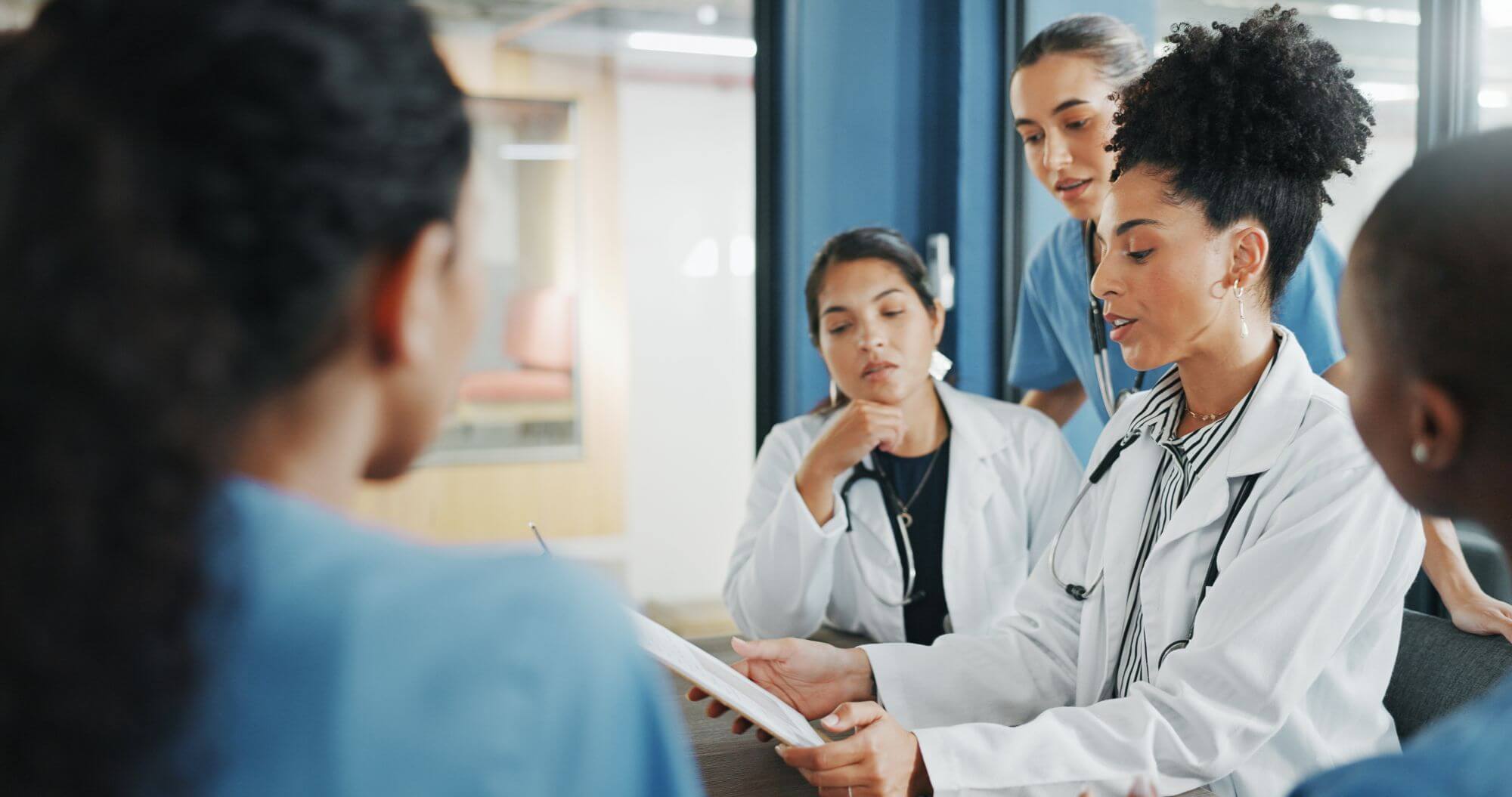 A diverse group of medical professionals in white coats and scrubs are gathered around a table discussing a document.
